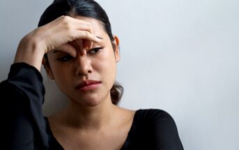 A woman with dark hair is leaning against a white wall, looking pensive. She is wearing a black top and has her right hand resting on her forehead. Her expression appears contemplative and slightly troubled.