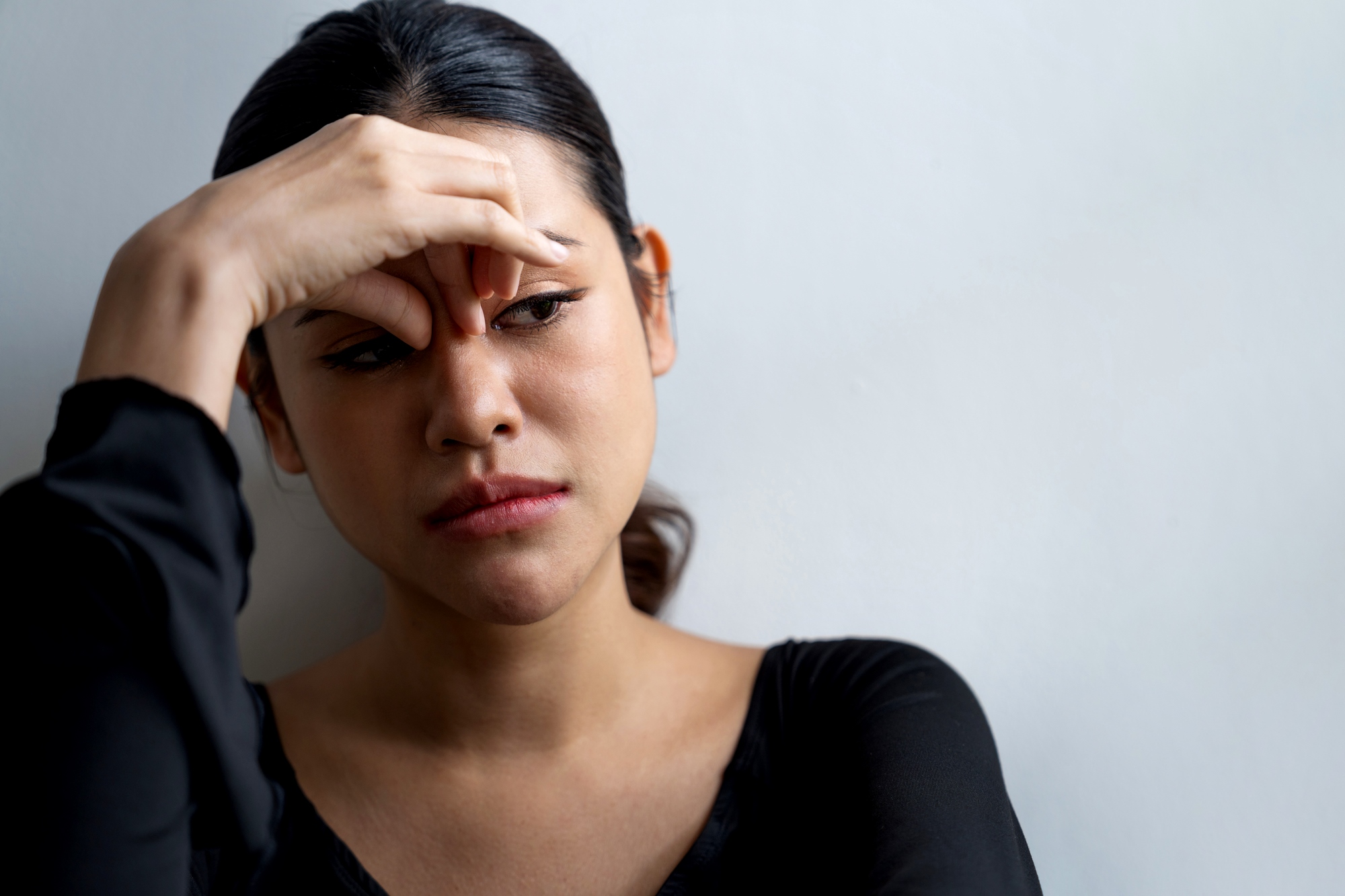 A woman with dark hair is leaning against a white wall, looking pensive. She is wearing a black top and has her right hand resting on her forehead. Her expression appears contemplative and slightly troubled.