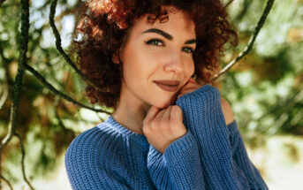 A person with curly hair smiles softly while posing outdoors. They are wearing a blue knit sweater, with their hands gently touching their face. Green foliage in the background gives the photo a serene, nature-like atmosphere.