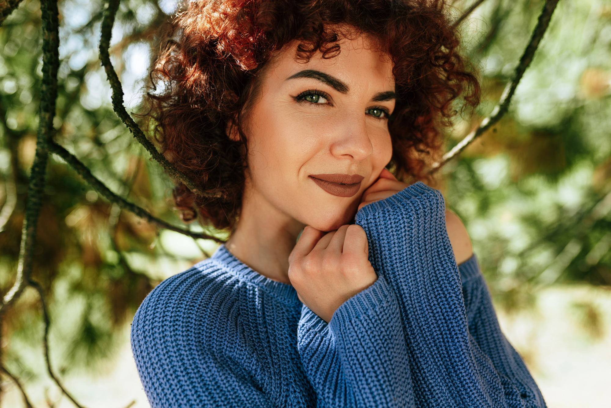 A person with curly hair smiles softly while posing outdoors. They are wearing a blue knit sweater, with their hands gently touching their face. Green foliage in the background gives the photo a serene, nature-like atmosphere.