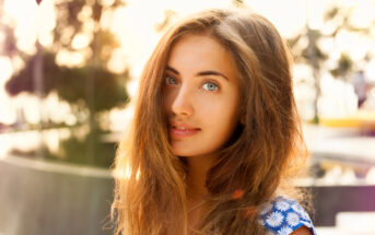 A young woman with long, light brown hair looks into the camera with a soft smile. She is outdoors, with sunlight creating a warm glow around her. She is wearing a blue top with white daisies and there are blurred trees and structures in the background.