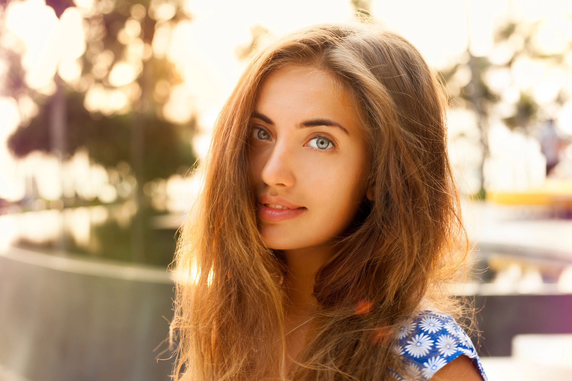 A young woman with long, light brown hair looks into the camera with a soft smile. She is outdoors, with sunlight creating a warm glow around her. She is wearing a blue top with white daisies and there are blurred trees and structures in the background.