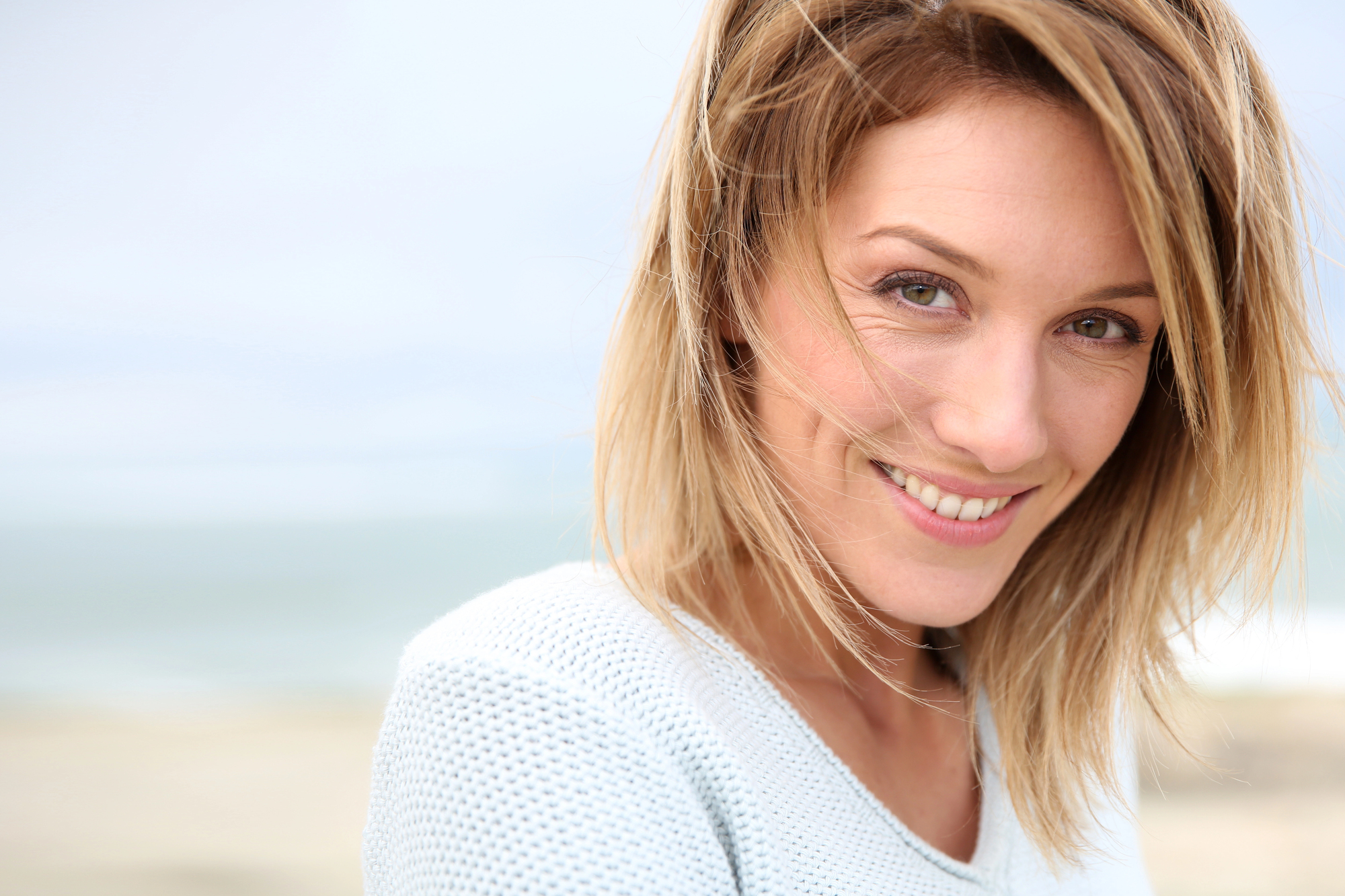 A woman with shoulder-length blonde hair and a light sweater smiles directly at the camera. The background is a blurred outdoor scene, suggesting a beach or coastal area. The sky is overcast, giving a soft and serene ambiance to the image.