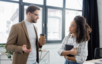 A man wearing glasses, a beige blazer, and a white turtleneck holds a coffee cup while talking to a woman with curly hair, wearing a plaid shirt and holding a black notebook. They are standing in a modern office with large windows in the background.