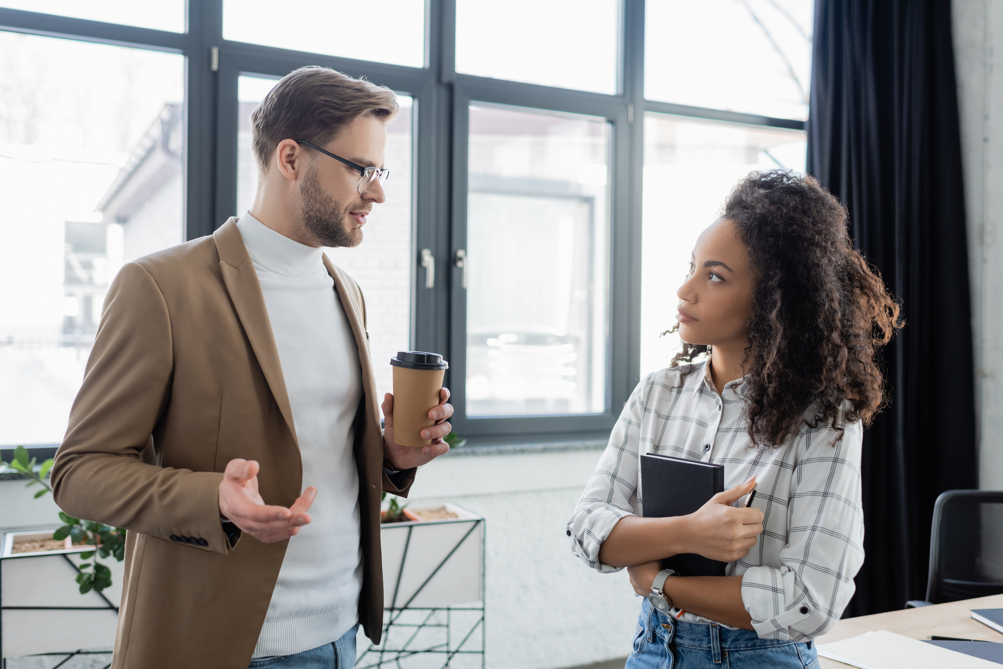 A man wearing glasses, a beige blazer, and a white turtleneck holds a coffee cup while talking to a woman with curly hair, wearing a plaid shirt and holding a black notebook. They are standing in a modern office with large windows in the background.