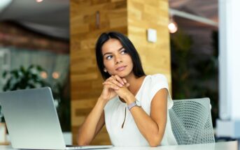 A woman with long dark hair sits at a desk in an office, looking thoughtfully to the side. She is wearing a white blouse and has her hands clasped under her chin. In front of her, there's an open laptop. The background is modern with wooden elements and plants.