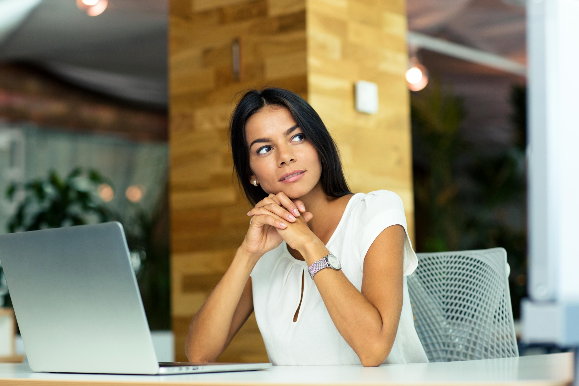 A woman with long dark hair sits at a desk in an office, looking thoughtfully to the side. She is wearing a white blouse and has her hands clasped under her chin. In front of her, there's an open laptop. The background is modern with wooden elements and plants.