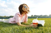 A woman wearing sunglasses lies on her stomach on a grassy field, holding an open book in one hand and a plastic cup with a straw filled with a colorful drink in the other. The sky is partly cloudy, and trees are visible in the background.