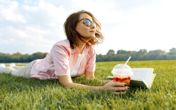 A woman wearing sunglasses lies on her stomach on a grassy field, holding an open book in one hand and a plastic cup with a straw filled with a colorful drink in the other. The sky is partly cloudy, and trees are visible in the background.