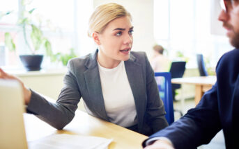 A woman with blonde hair, wearing a gray blazer and white shirt, sits at a desk and talks to a man in a suit. She gestures with one hand, possibly explaining something. They are in a bright office with large windows and several potted plants in the background.