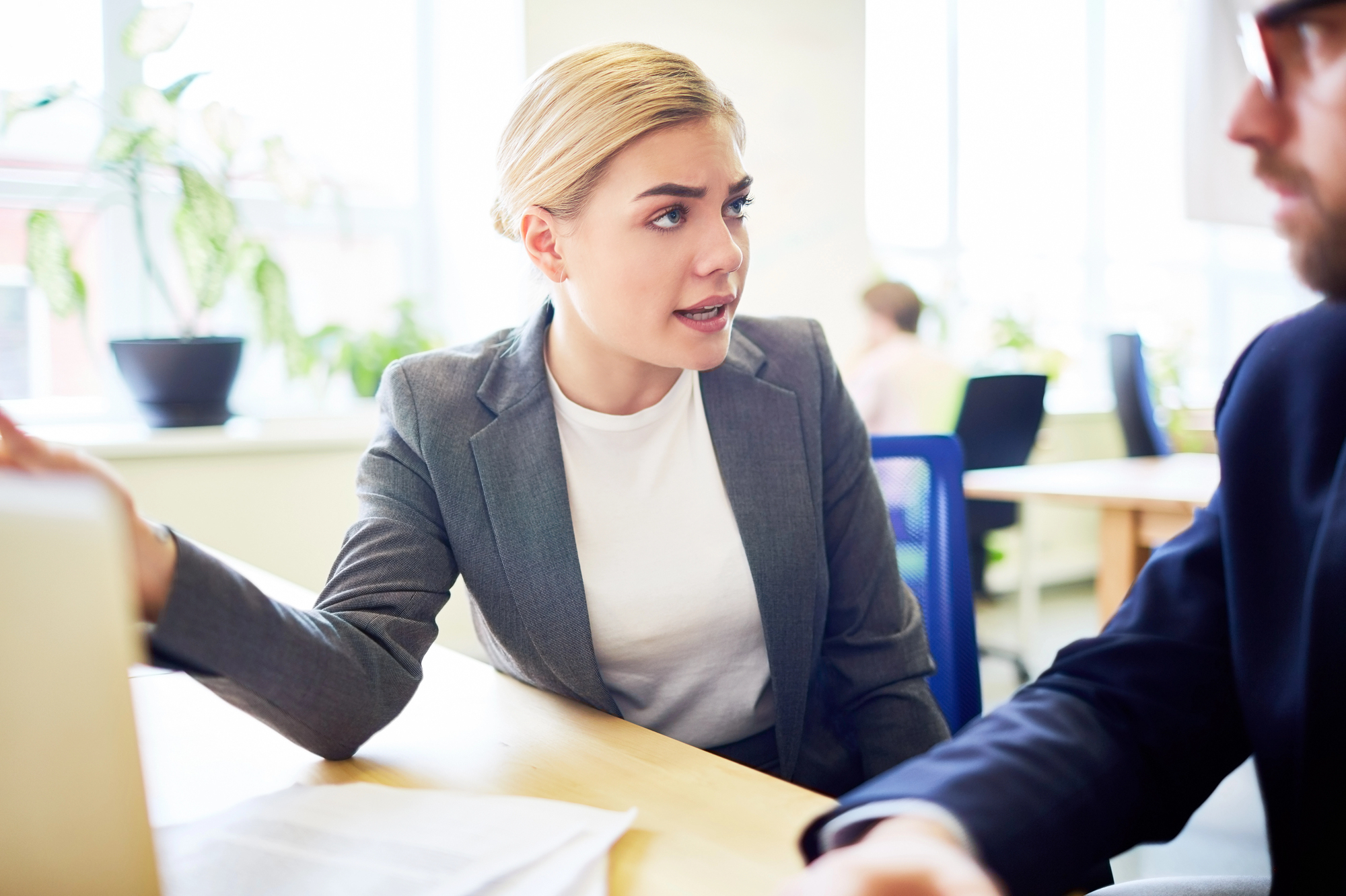 A woman with blonde hair, wearing a gray blazer and white shirt, sits at a desk and talks to a man in a suit. She gestures with one hand, possibly explaining something. They are in a bright office with large windows and several potted plants in the background.