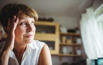 A woman with short hair and a white sleeveless blouse gazes thoughtfully out of a window. She has one hand resting on her temple. The background shows a kitchen with shelves containing various items. Soft natural light illuminates the scene.