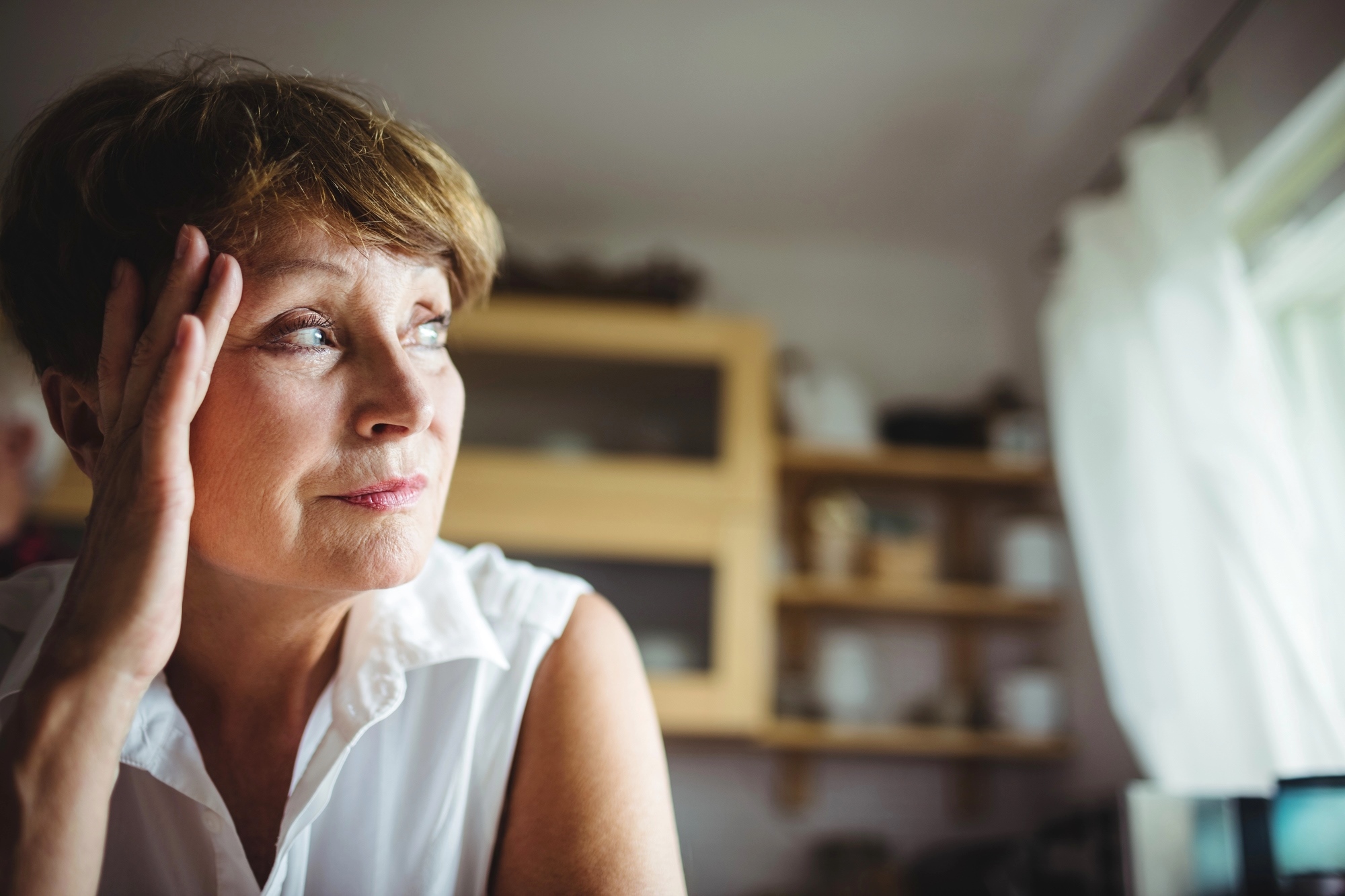 A woman with short hair and a white sleeveless blouse gazes thoughtfully out of a window. She has one hand resting on her temple. The background shows a kitchen with shelves containing various items. Soft natural light illuminates the scene.