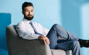 A man with a beard and mustache, dressed in a white shirt, blue tie, and blue pants, is sitting confidently on a gray chair against a light blue background. His legs are crossed, and he is looking directly at the camera.