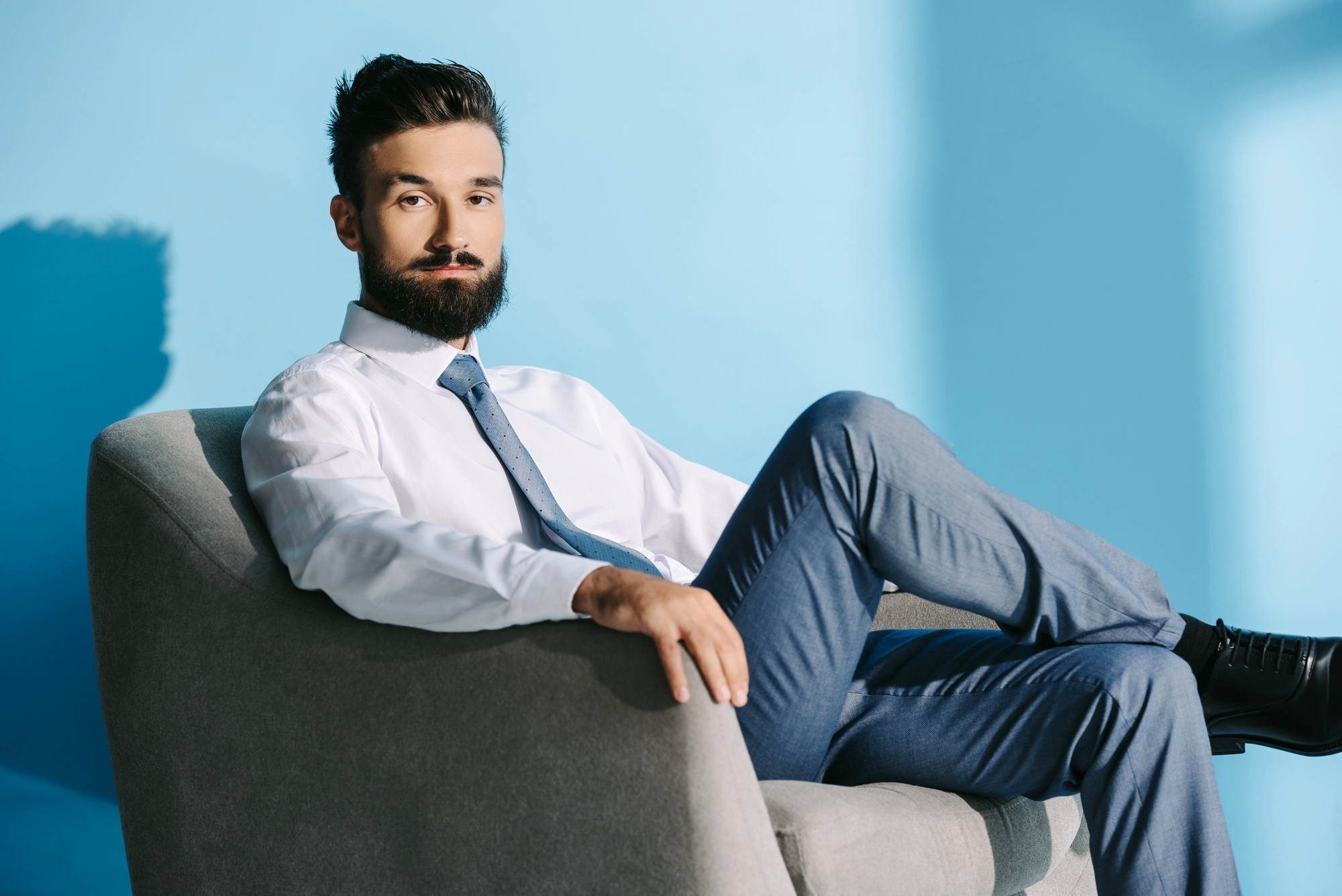 A man with a beard and mustache, dressed in a white shirt, blue tie, and blue pants, is sitting confidently on a gray chair against a light blue background. His legs are crossed, and he is looking directly at the camera.