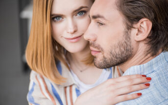 A close-up of a woman with shoulder-length blonde hair smiling at the camera while hugging a man with short brown hair and a trimmed beard, who faces sideways. The woman wears a striped shirt, and both appear to be in a casual indoor setting.