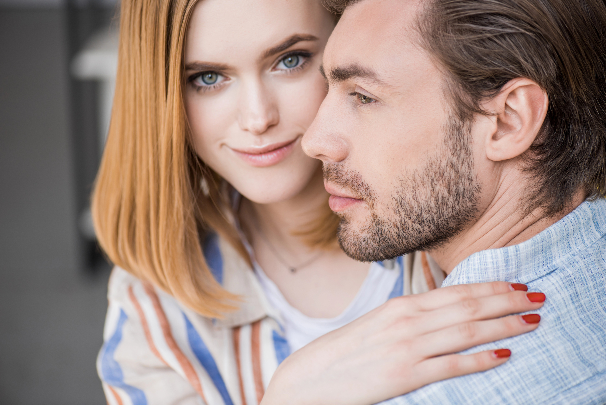 A close-up of a woman with shoulder-length blonde hair smiling at the camera while hugging a man with short brown hair and a trimmed beard, who faces sideways. The woman wears a striped shirt, and both appear to be in a casual indoor setting.