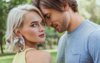 A woman with short blonde hair and large floral earrings looks at the camera, while a man with medium-length brown hair in a blue striped shirt leans his forehead against hers. They are outside, with greenery in the background.