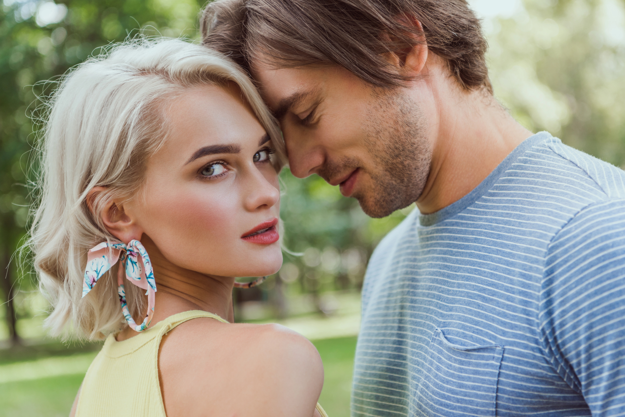 A woman with short blonde hair and large floral earrings looks at the camera, while a man with medium-length brown hair in a blue striped shirt leans his forehead against hers. They are outside, with greenery in the background.