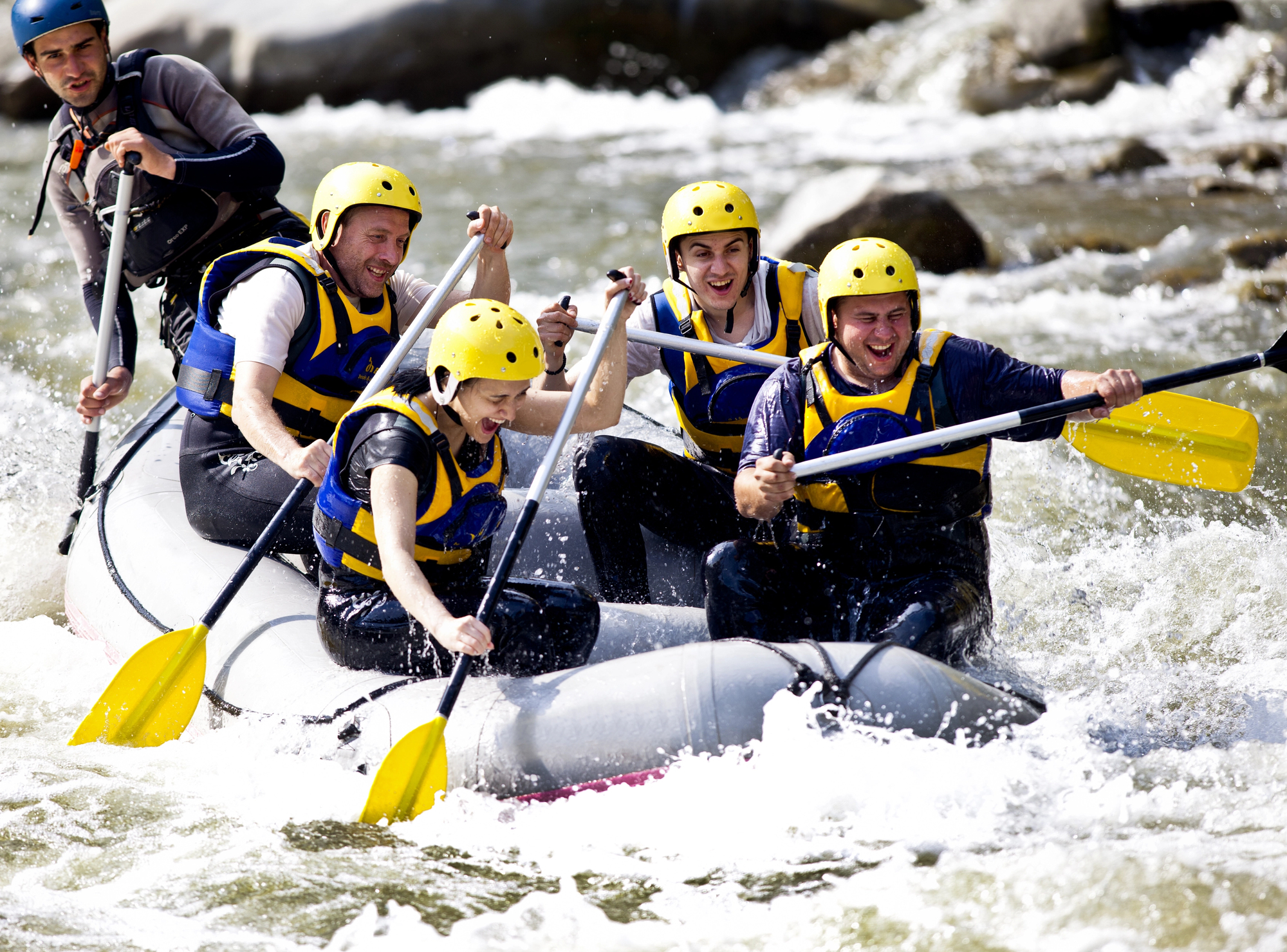 Five people wearing yellow helmets and life jackets paddle vigorously in a white water raft, navigating through a rapid in a river. The water splashes around them as they work together to steer the inflatable raft.