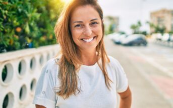 A woman with long, light brown hair smiles brightly at the camera while standing outdoors on a sunny day. She is wearing a white short-sleeve shirt and has one hand resting on her hip. A street with parked cars and greenery is visible in the background.