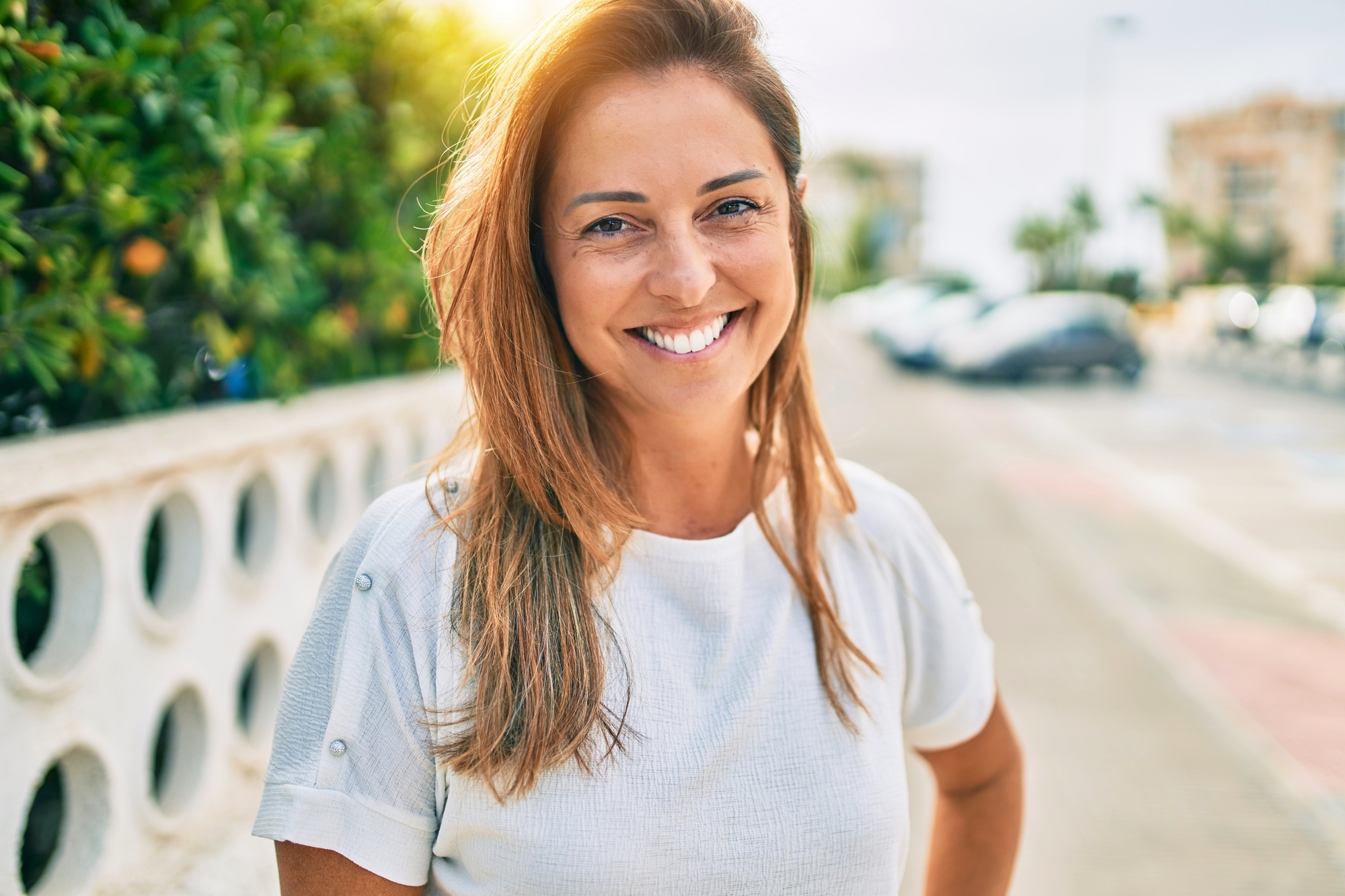 A woman with long, light brown hair smiles brightly at the camera while standing outdoors on a sunny day. She is wearing a white short-sleeve shirt and has one hand resting on her hip. A street with parked cars and greenery is visible in the background.