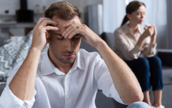 A man sits on the floor with his head in his hands, appearing stressed or upset. A woman sits on a couch in the background, looking contemplative and not facing him. The scene depicts tension or conflict in a domestic setting.