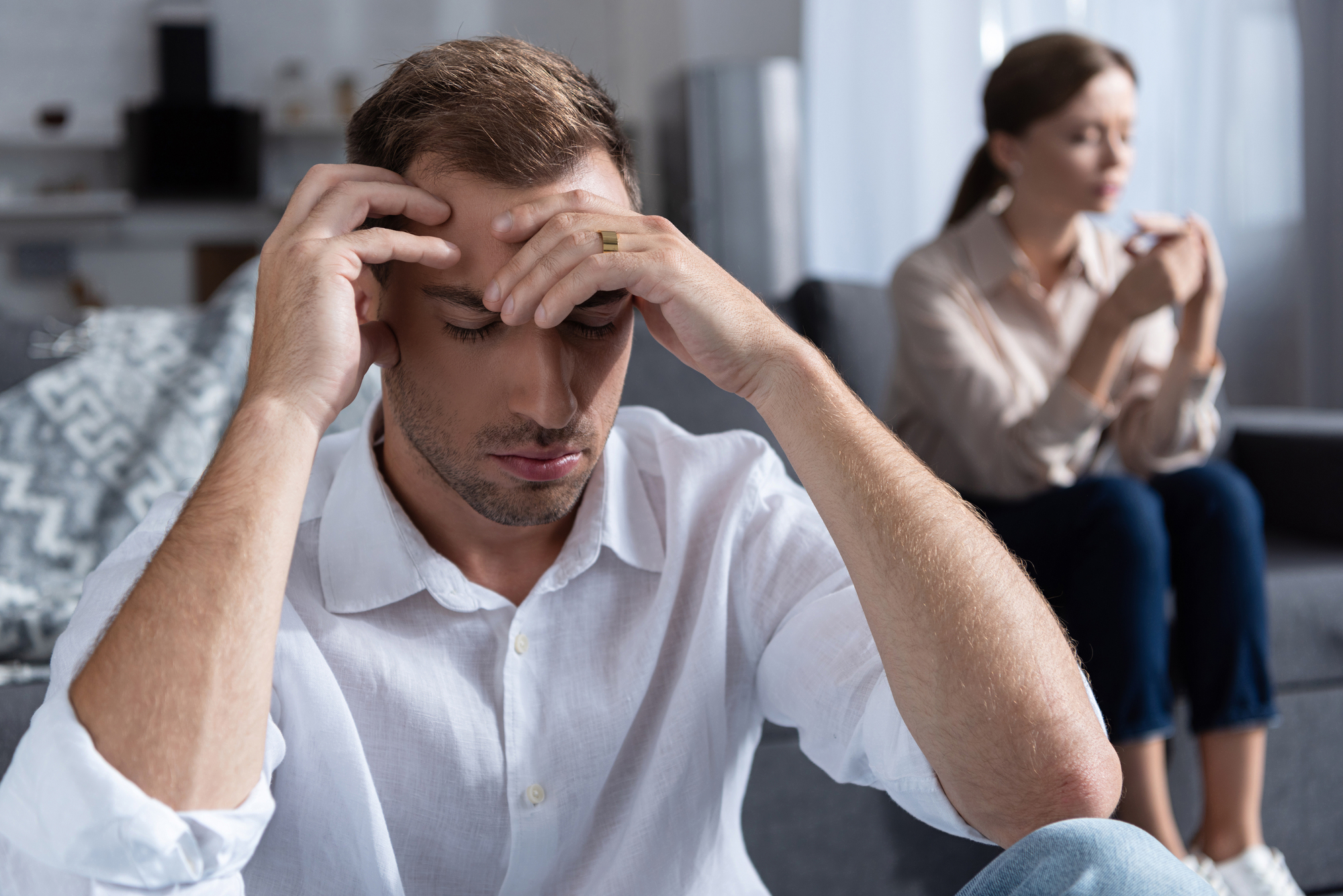 A man sits on the floor with his head in his hands, appearing stressed or upset. A woman sits on a couch in the background, looking contemplative and not facing him. The scene depicts tension or conflict in a domestic setting.