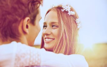 A woman with long hair, adorned with a floral headband, smiles brightly at a man facing her. The scene is outdoors with a soft, golden sunlight in the background, creating a warm and romantic atmosphere.