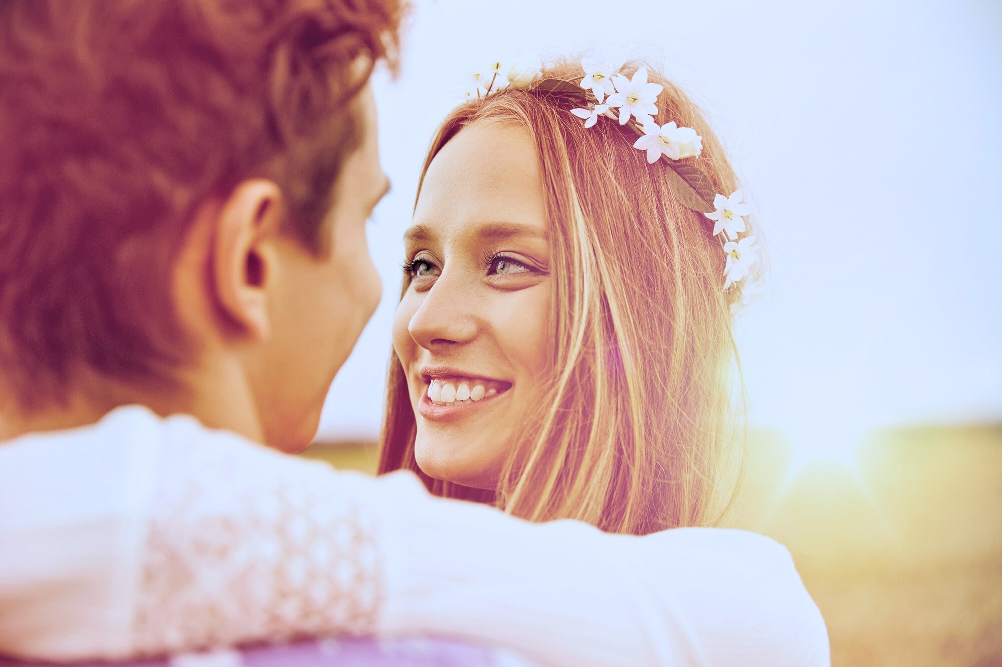 A woman with long hair, adorned with a floral headband, smiles brightly at a man facing her. The scene is outdoors with a soft, golden sunlight in the background, creating a warm and romantic atmosphere.