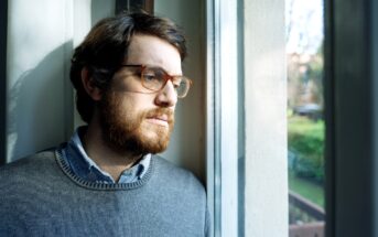 A man with glasses and a beard is standing by a window, looking outside. He is wearing a gray sweater over a collared shirt. Natural light is illuminating his face, creating a thoughtful expression. The background shows a blurred view of trees and outdoor scenery.