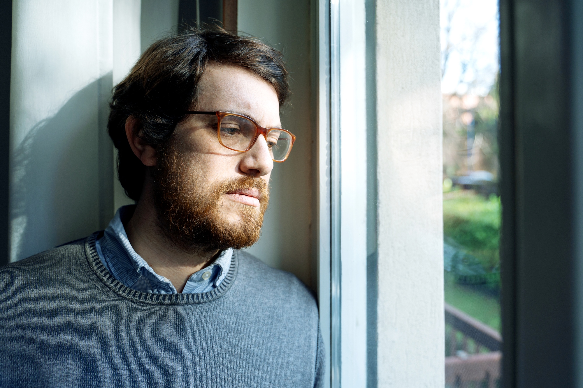 A man with glasses and a beard is standing by a window, looking outside. He is wearing a gray sweater over a collared shirt. Natural light is illuminating his face, creating a thoughtful expression. The background shows a blurred view of trees and outdoor scenery.