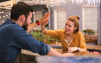 A man and a woman high-five each other in a bar. The woman, wearing a yellow blouse, is holding a martini glass with an olive. The man, in a blue shirt and apron, is smiling and reaching out. Wine glasses hang overhead, and warm lighting creates a cozy atmosphere.