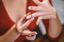 A person with white polished nails and a red top is slipping a gold ring onto their ring finger. Their other hand supports and guides the ring. The scene appears to be intimate and focused on the act of putting on the ring.