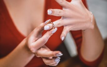 A person with white polished nails and a red top is slipping a gold ring onto their ring finger. Their other hand supports and guides the ring. The scene appears to be intimate and focused on the act of putting on the ring.