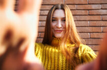 A person with long hair stands in front of a brick wall, smiling at the camera. They are wearing a bright yellow knitted sweater, and their hands are partially visible in the foreground, reaching towards the camera.