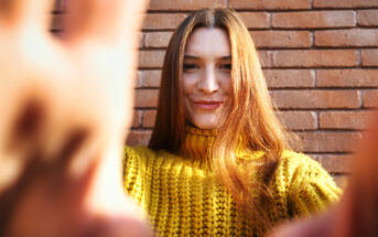 A person with long hair stands in front of a brick wall, smiling at the camera. They are wearing a bright yellow knitted sweater, and their hands are partially visible in the foreground, reaching towards the camera.