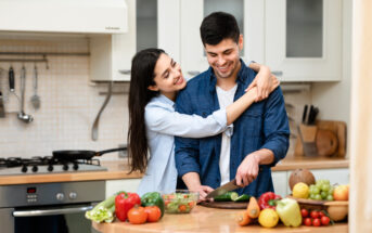 A smiling couple is in a modern kitchen. The man is chopping vegetables on a wooden board while the woman hugs him from behind. The countertop is filled with various colorful fresh fruits and vegetables. They appear to be sharing a joyful moment together.