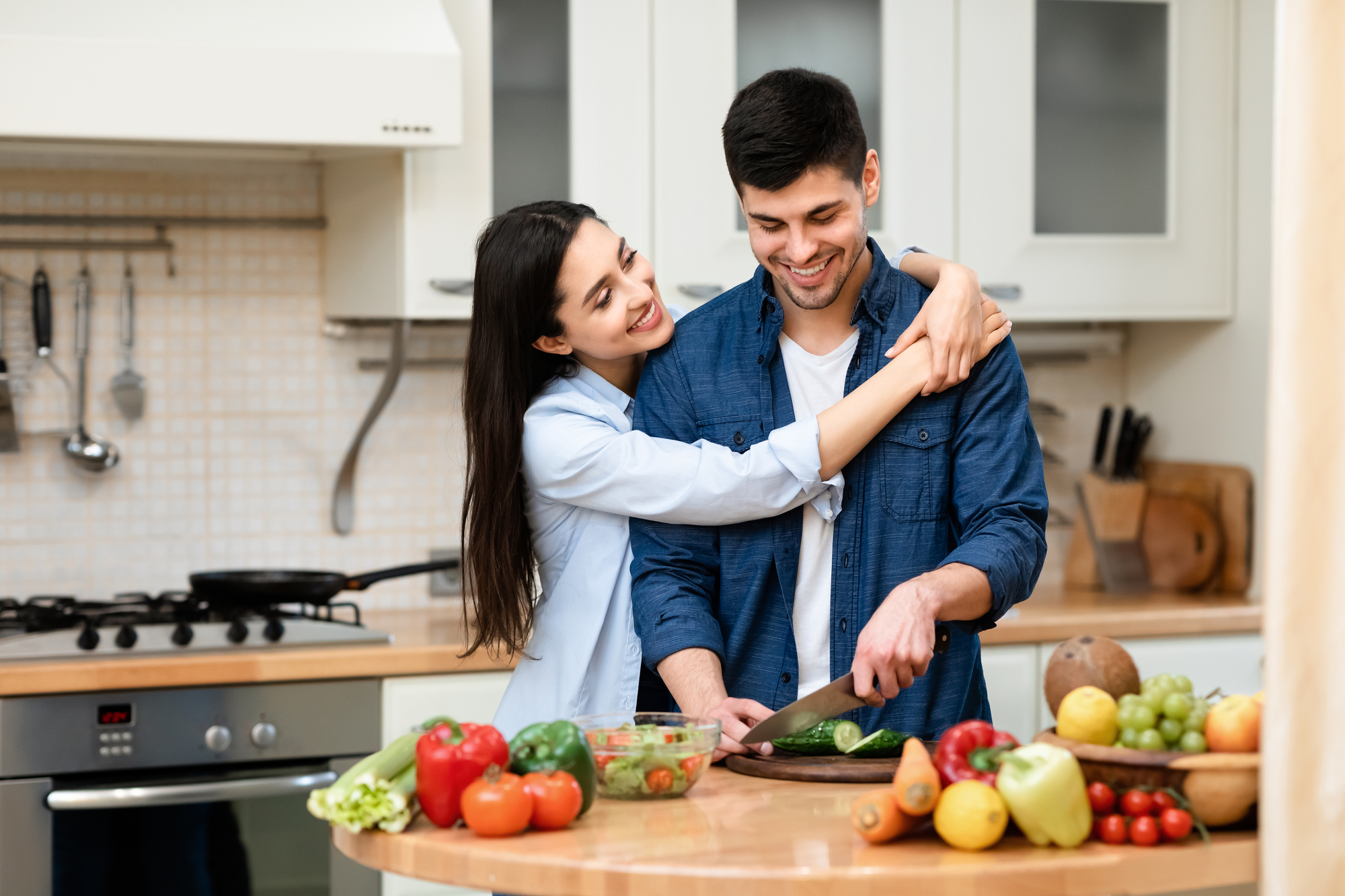 A smiling couple is in a modern kitchen. The man is chopping vegetables on a wooden board while the woman hugs him from behind. The countertop is filled with various colorful fresh fruits and vegetables. They appear to be sharing a joyful moment together.