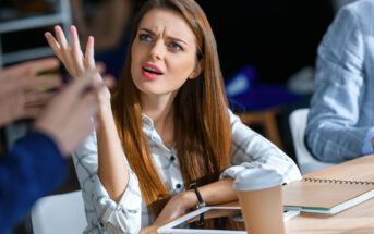 A woman with long brown hair and a plaid shirt sits at a table with a tablet, notebook, and coffee cup. She looks confused or frustrated, raising one hand in a questioning gesture while speaking to an unseen person. The background is out of focus.