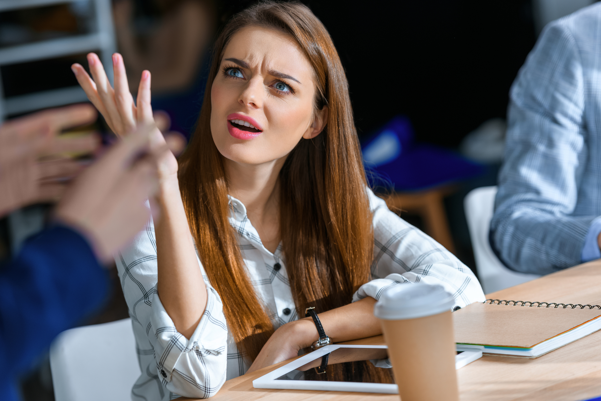 A woman with long brown hair and a plaid shirt sits at a table with a tablet, notebook, and coffee cup. She looks confused or frustrated, raising one hand in a questioning gesture while speaking to an unseen person. The background is out of focus.