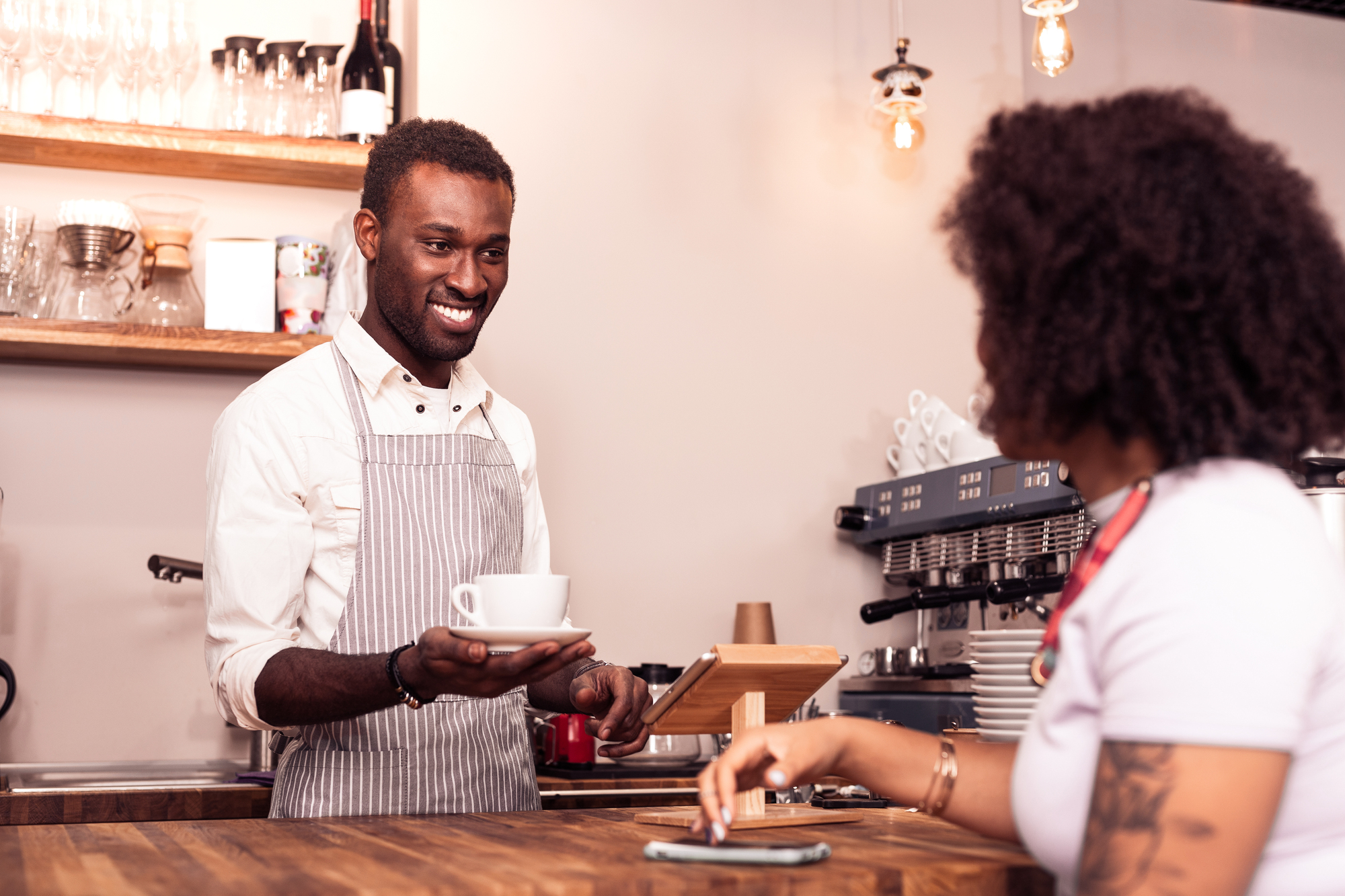 A barista, wearing a grey apron and white shirt, stands behind a counter and smiles while serving a cup of coffee to a customer with curly hair and a tattoo on her arm. The background features shelves with glasses and coffee equipment.