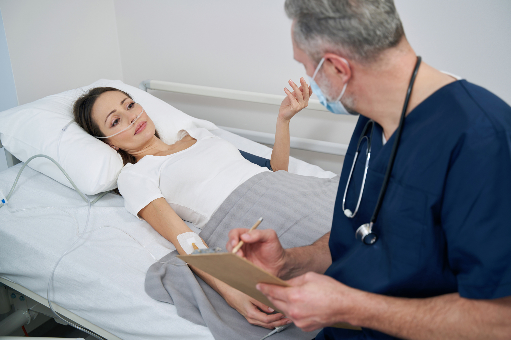 A woman lying in a hospital bed with a nasal cannula and a monitor attached to her arm is talking to a male healthcare professional in scrubs, who is holding a clipboard and writing notes. The healthcare professional is wearing a mask.