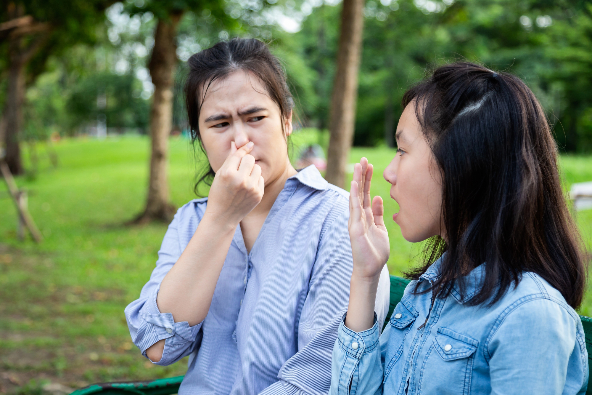 Two women are sitting outside in a park. One woman is holding her nose with a disgusted expression, while the other woman appears to be explaining something with her hand raised. They are surrounded by greenery with trees and grass in the background.