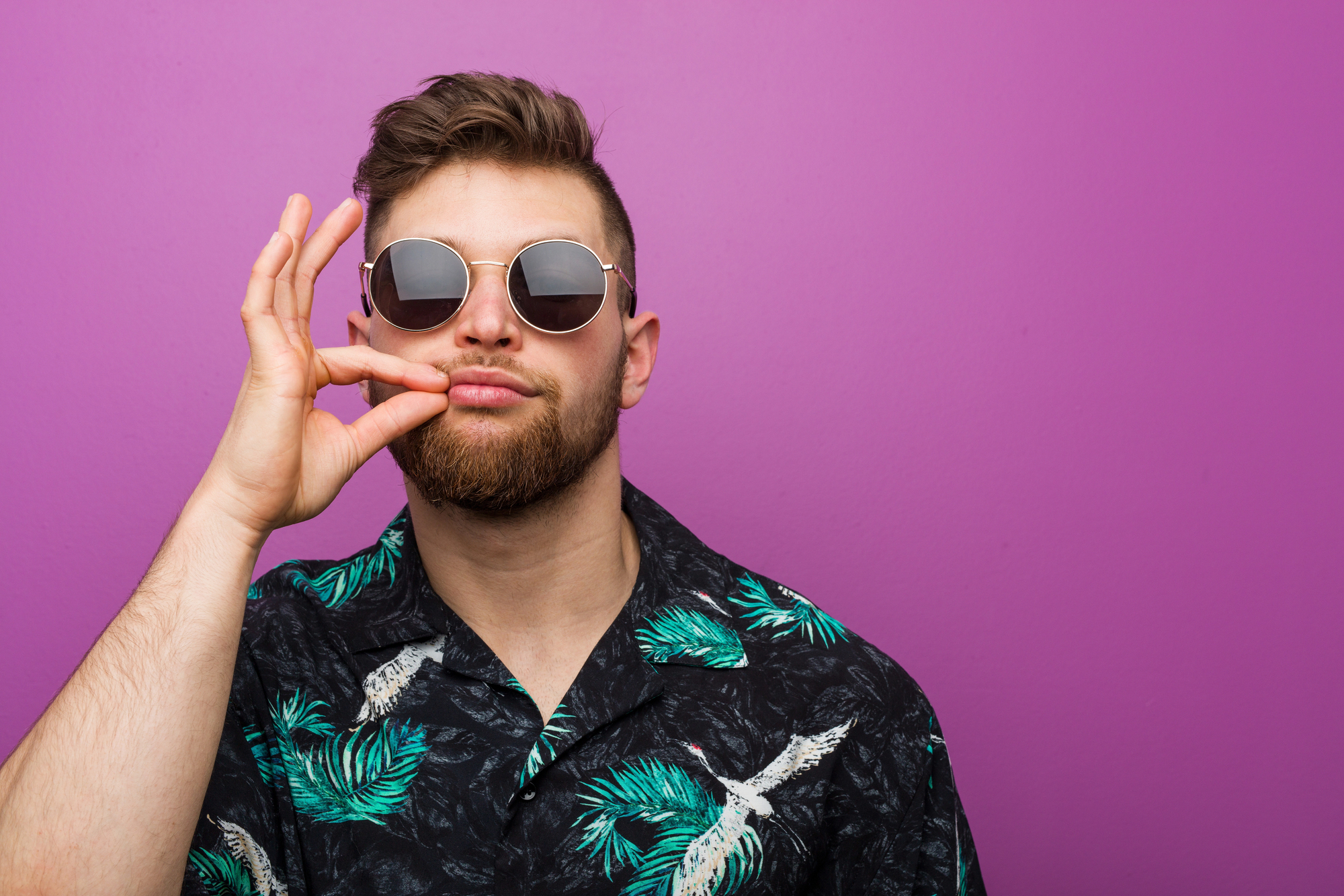 A bearded man with sunglasses and a black shirt featuring green palm leaf print poses in front of a purple background. He is making an OK sign with his right hand and holding it near his mouth.