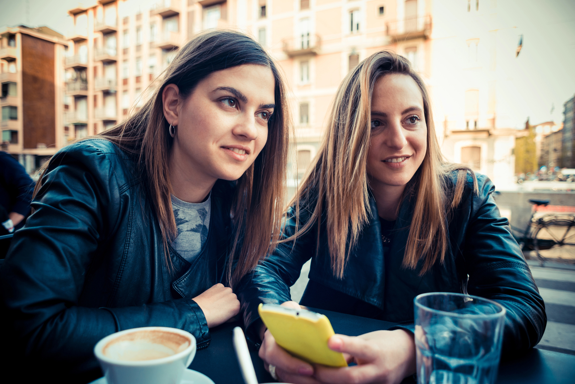 Two women sit at an outdoor café table with drinks in front of them. Both are wearing dark jackets and have long hair. One is holding a yellow smartphone while they both look in the same direction, appearing engaged in something off-camera. Buildings are in the background.