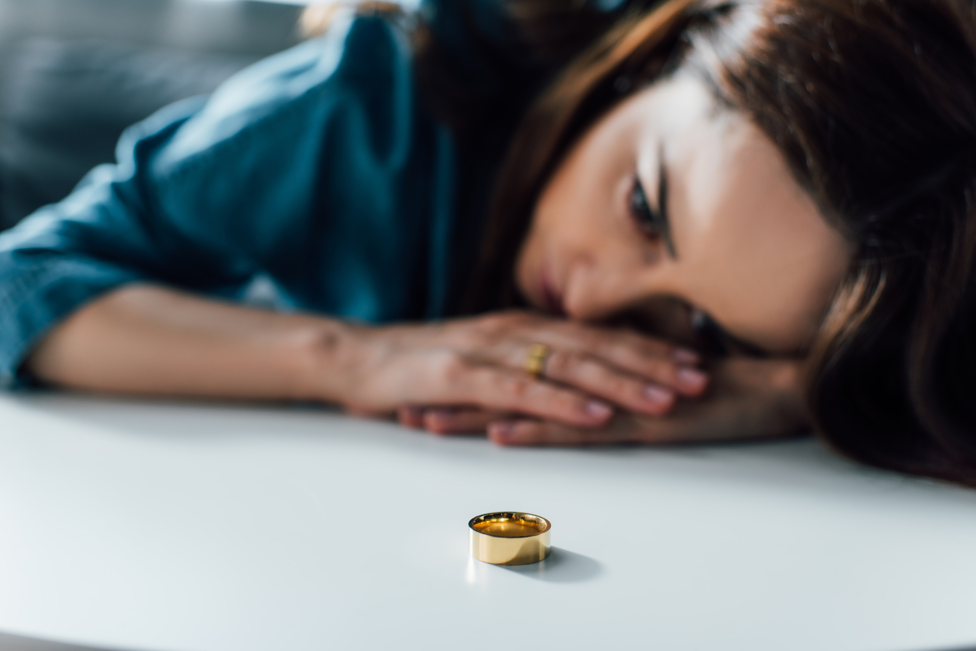 A woman with brunette hair lies on a table, her hands under her head, staring at a gold ring placed in front of her. She appears thoughtful or sad, with the focus on the ring in the foreground.