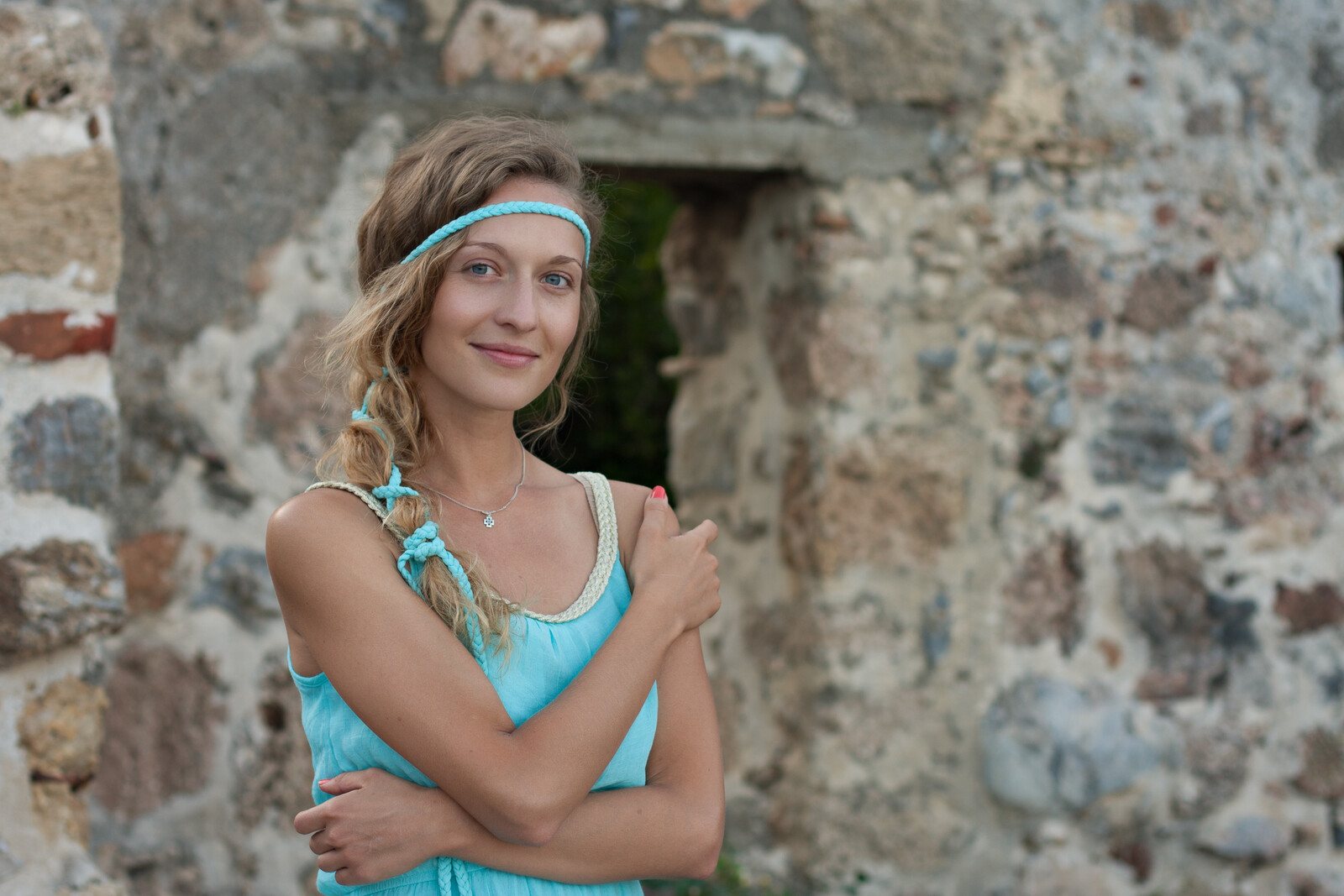 A woman with a braided hairstyle and headband stands in front of a weathered stone wall. She is wearing a turquoise dress and has her arms crossed with a faint smile on her face. The background features an arched doorway and a rustic stone texture.