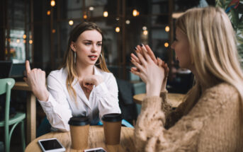 Two women are sitting at a wooden table in a cozy café, engaged in an animated conversation. Each has a takeaway coffee cup in front of them, along with their smartphones. One woman is gesturing with her hands, while the other listens attentively.