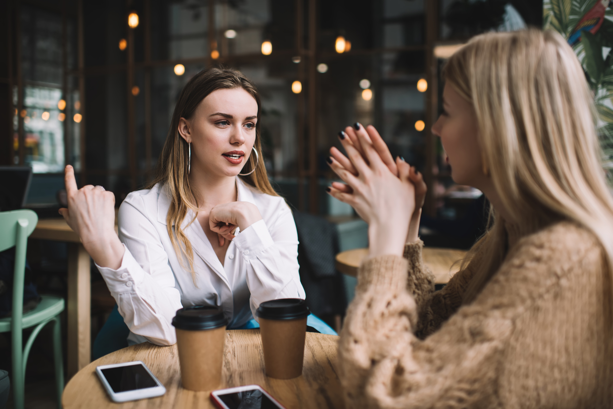 Two women are sitting at a wooden table in a cozy café, engaged in an animated conversation. Each has a takeaway coffee cup in front of them, along with their smartphones. One woman is gesturing with her hands, while the other listens attentively.